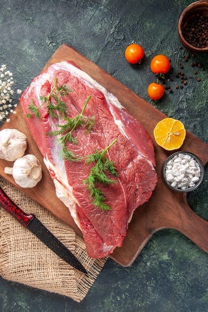 Overhead view of fresh raw red meats green garlics lemon salt on brown wooden cutting board knife on nude color towel tomatoes pepper on dark color background