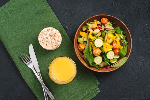 An overhead view of fresh vegetable salad; puffed rice cake and juice with cutlery on napkin over black concrete backdrop