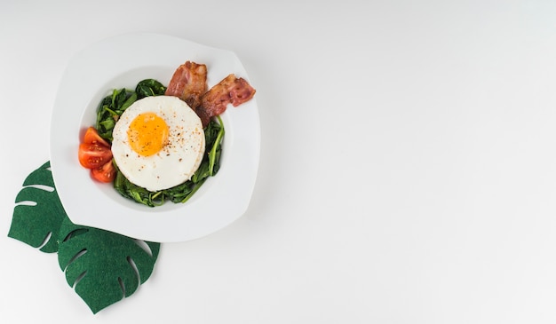 An overhead view of an fried egg with spinach; tomato and bacon on white plate against white background