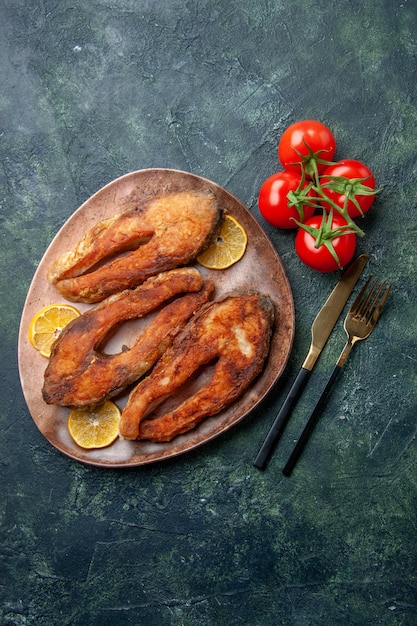 Overhead view of fried fishes and lemon slices on a brown plate tomatoes cutlery set on mix colors table with free space