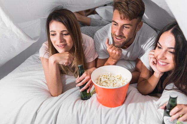 An overhead view of friends lying on bed enjoying drink and popcorn