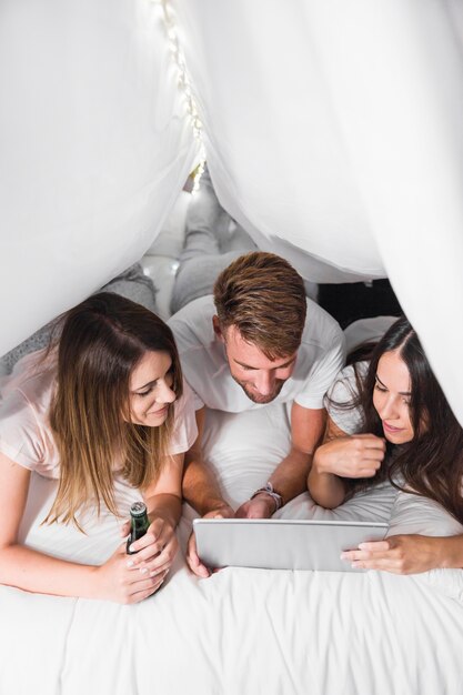 An overhead view of friends lying on white bed looking at digital tablet