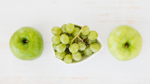 An overhead view of green apples with grapes on white wooden textured backdrop