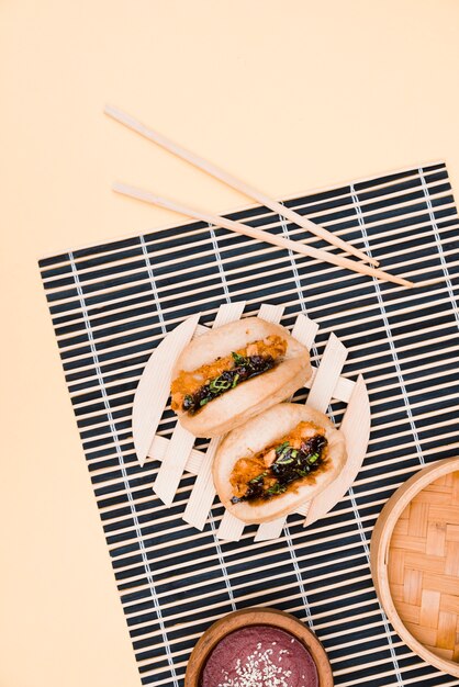 An overhead view of gua bao meat and chicken asian food on placemat with chopsticks against beige backdrop
