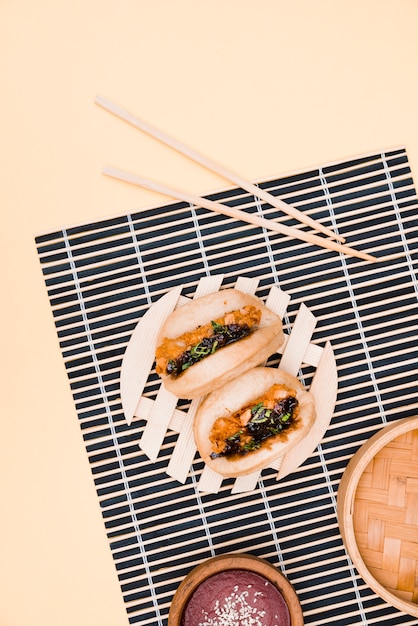 An overhead view of gua bao meat and chicken asian food on placemat with chopsticks against beige backdrop