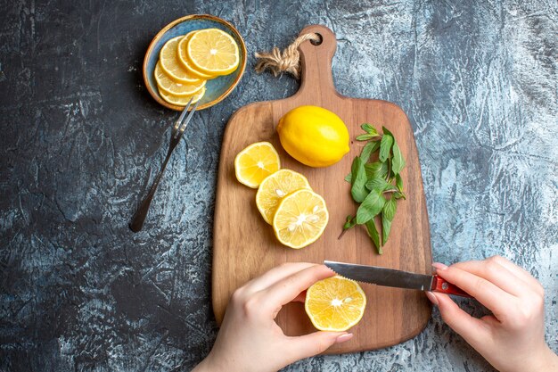 Overhead view of a hand chopping fresh lemons and mint on a wooden cutting board on dark background