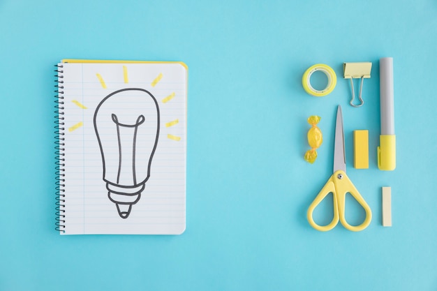 Overhead view of hand drawn light bulb on notebook with stationary on blue background