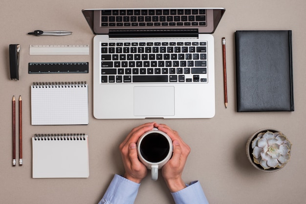 An overhead view of hand holding coffee cup with office supplies and laptop on colored desk