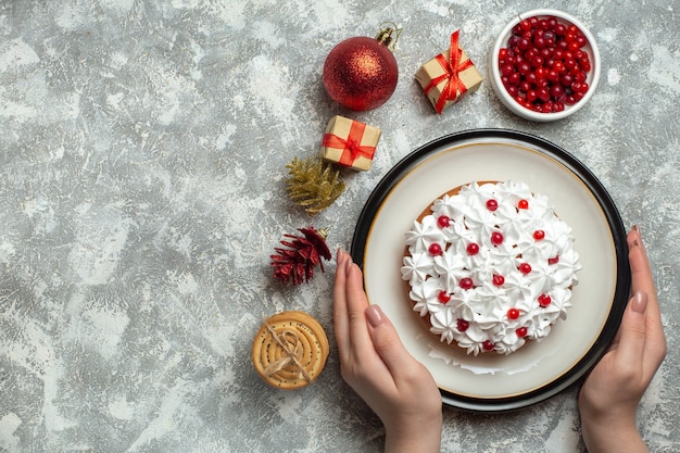 Overhead view of hand holding delicious cake with cream currant on a plate and gift boxes stacked cookies conifer cones on gray background