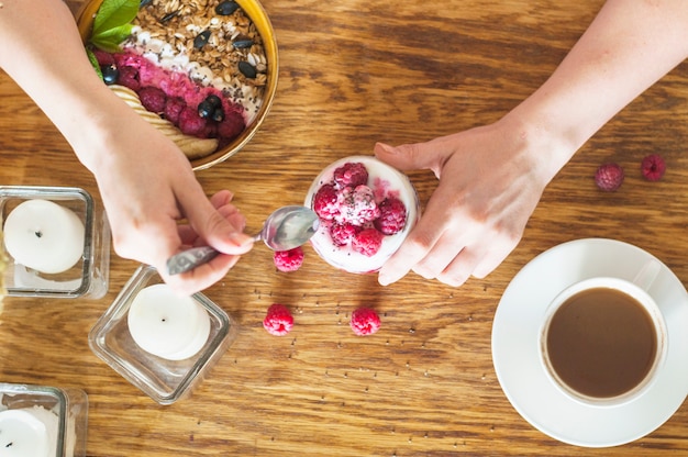 Верхний вид руки, держащей ложку и стакан йогурта с малиной на деревянном столе