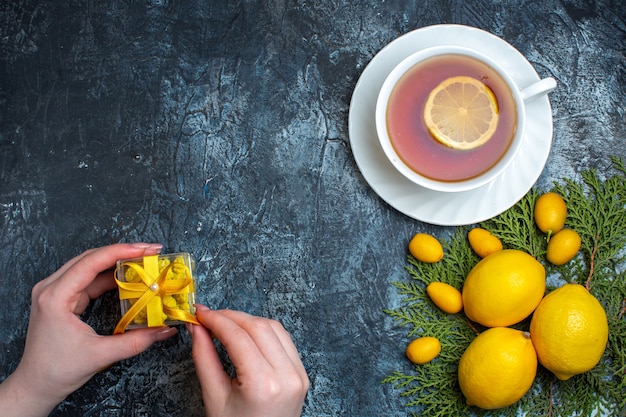 Overhead view of hand opening a gift and a cup of black tea with lemon next to citrus fruits collection on fir branches on dark background