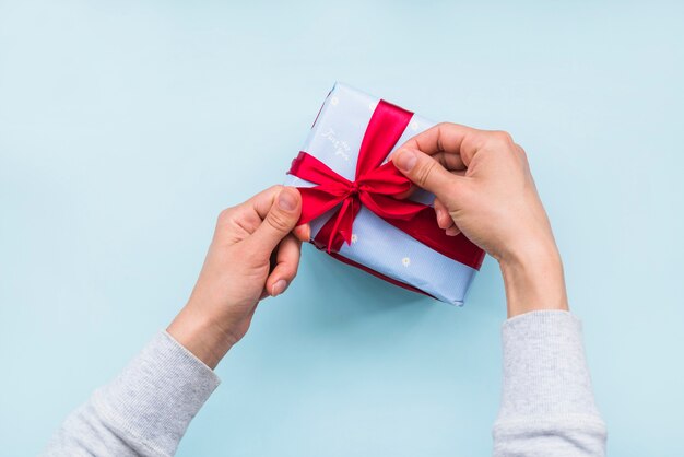 Overhead view of hand tying red ribbon bow on gift box over blue background