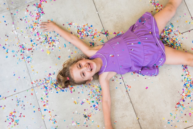 Overhead view of happy cute girl lying with confetti on floor