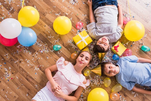 Overhead view of happy siblings lying with balloons; gift box and confetti on floor