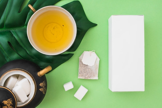 Верхний вид травяной чашки чая с кубиками сахара, чайный пакетик и коробка на зеленом фоне