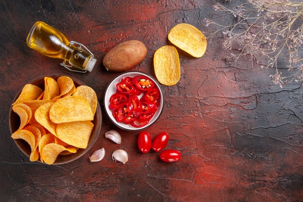 Overhead view of homemade delicious crispy potato chips in a brown pot fallen oil bottle ketchup tomatoes potato garlic on dark background