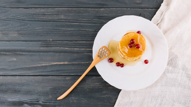 An overhead view of homemade pancakes with honey and berries on plate against wooden plank