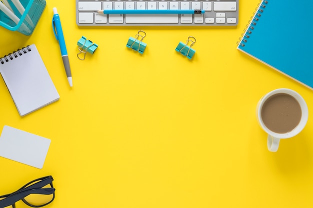 An overhead view of keyboard; eyeglasses and tea cup on yellow workspace