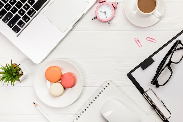 An overhead view of laptop,alarm clock,coffee cup,macaroons,pencil,mouse,spiral notepad on white desk