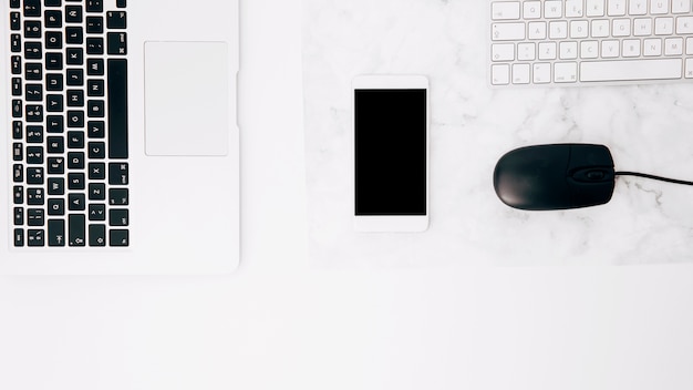 An overhead view of laptop; cellphone; mouse and keyboard on white desk