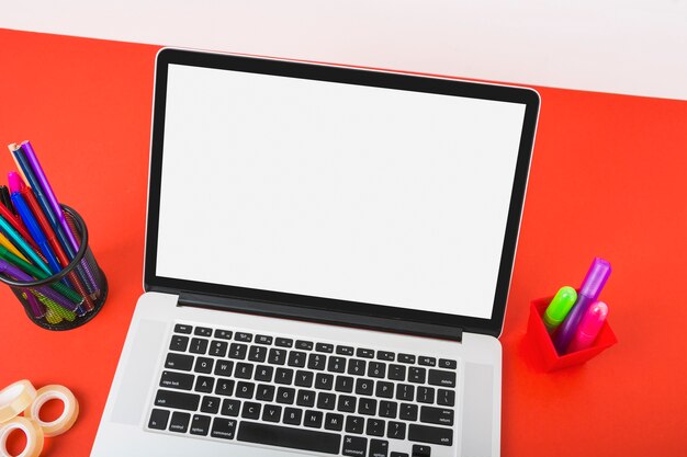 An overhead view of laptop displaying white screen with colorful stationeries on red desk