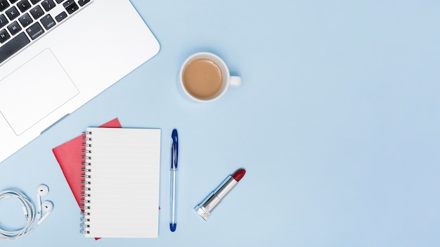 Overhead view of laptop; earphones; blank notepad; pen; lipstick and cup of a tea on blue background