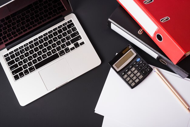 An overhead view of laptop; file folders; calculator; pencils and paper on black background