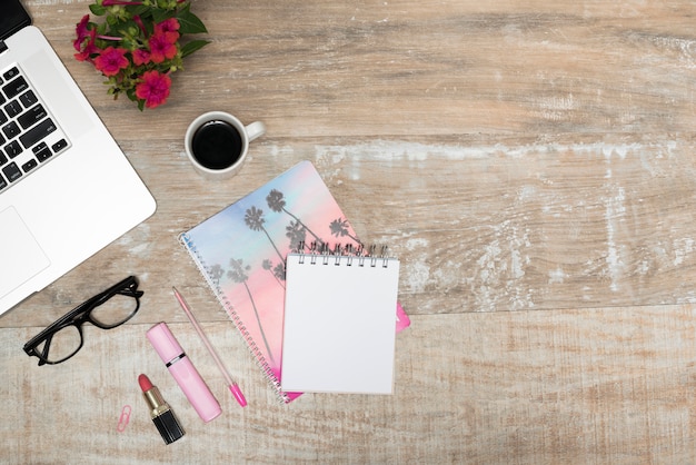 Overhead view of laptop; lipstick; highlighter; spiral notepad; coffee cup; pen; and eyeglass on wooden office table