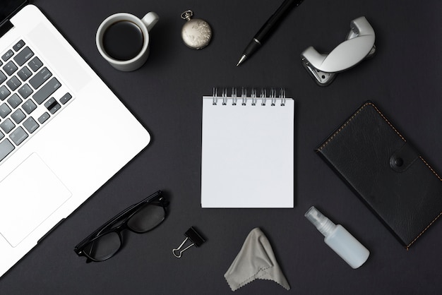 Overhead view of laptop and office stationery; coffee cup; eyeglasses; pen against black desktop