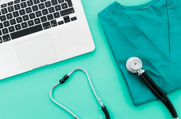 An overhead view of laptop; stethoscope and medical uniform on green backdrop