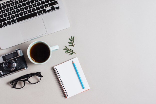 An overhead view of laptop; vintage camera; eyeglasses; tea cup and colored pencil on notepad against white backdrop