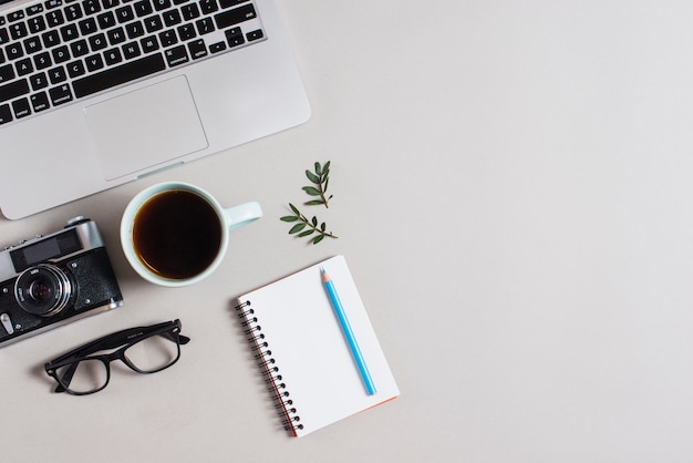 An overhead view of laptop; vintage camera; eyeglasses; tea cup and colored pencil on notepad against white backdrop