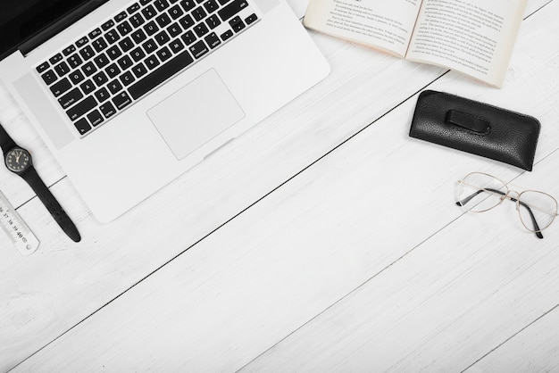 An overhead view of laptop with book; eyeglasses; ruler and wrist watch on white wooden table