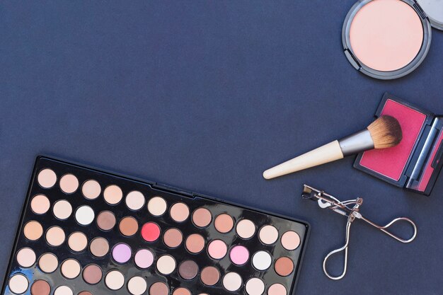 An overhead view of makeup brush; blusher; eyeshadow palette and eyelash curler on blue backdrop