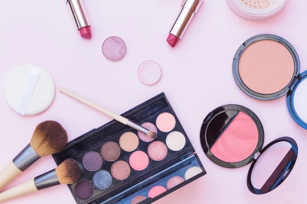 Overhead view of makeup brush; lipstick; eye shadow; sponge and blusher on pink background