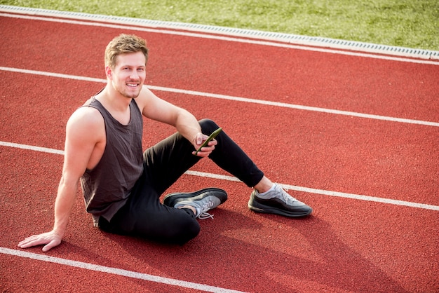 An overhead view of male athlete sitting on red race track holding mobile phone in hand