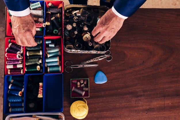 An overhead view of male fashion designer's hand holding thread spool and buttons from the box on wooden desk