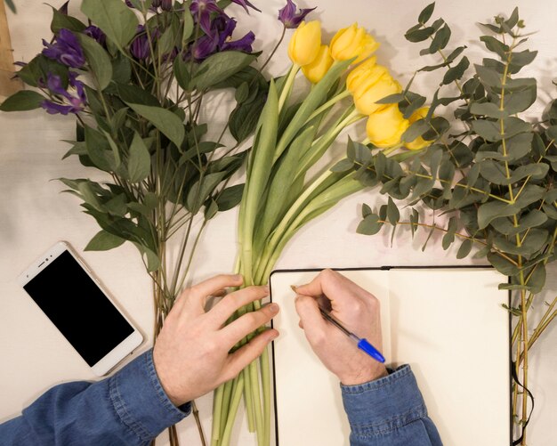 An overhead view of a male florist writing on diary with pen on table with flowers