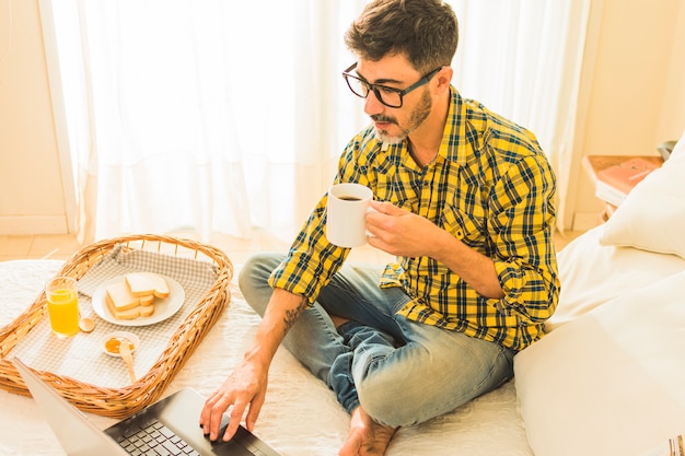 An overhead view of a man sitting on bed holding cup of coffee using laptop