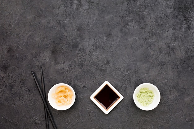 An overhead view of marinated ginger; wasabi and soya sauce with chopstick over blackboard