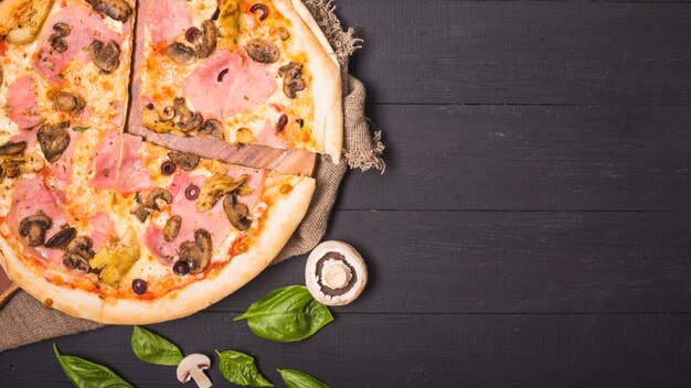 An overhead view of meat pizza with mushroom and basil leaf on wooden plank