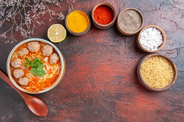 Overhead view of meatballs soup with noodles in a brown bowl lemon spoon and uncooked pasta and different spices on dark table