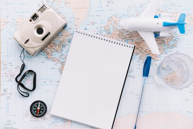 An overhead view of miniature white airplane; spiral blank notepad; pen; camera and compass on map