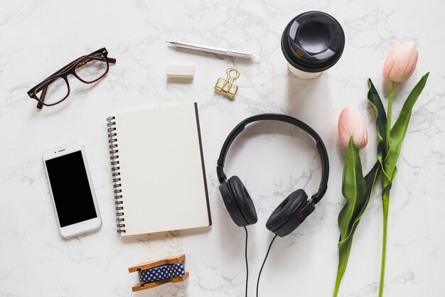 Overhead view of mobile phone; eyeglasses; notebook; stationery; headphone and pink tulips on marble backdrop