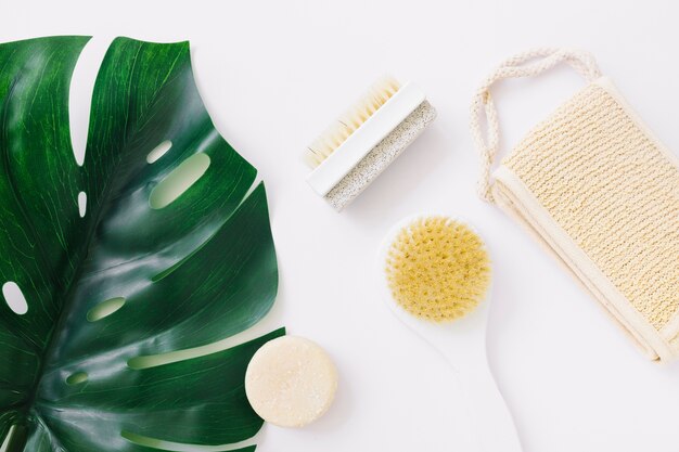 An overhead view of monstera leaf with loofah; soap; brush and pumice stone brush over white backdrop