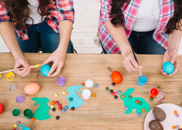 An overhead view of mother and daughter painting the easter eggs over the wooden table