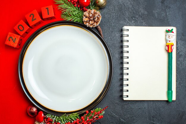 Overhead view of New year background with dinner plate decoration accessories fir branches and numbers on a red napkin next to notebook with pen on a black table