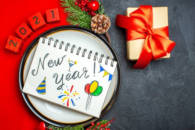 Overhead view of New year background with notebook with new year drawings on a dinner plate decoration accessories fir branches and numbers on a red napkin and a gift on a black table