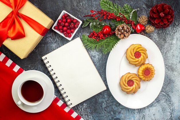 Overhead view of notebook a cup of black tea on a red towel and biscuits on a white plate new year accessories gift with red ribbon on dark surface