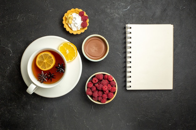 Overhead view of notebook and a cup of black tea with lemon served with chocolate raspberry honey on dark background